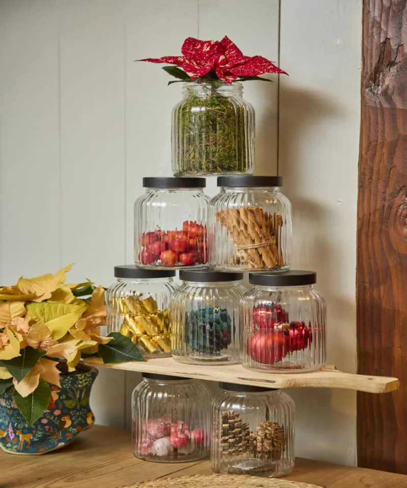 Świąteczna dekoracja z przezroczystych słoików ułożonych w formie choinki, z ozdobami i ciętą poinsecją na szczycie