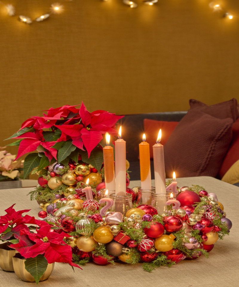 Kolorowy wieniec adwentowy z bombek i świec, ozdobiony czerwoną poinsecją na świątecznie udekorowanym stole