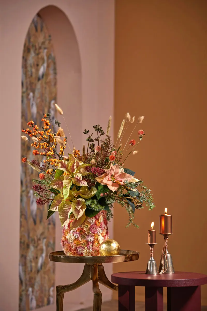 Bukiet z pastelowych poinsecji, gałązek i suszonych roślin w ozdobnym wazonie, obok świec w miedzianych świecznikach
