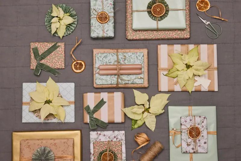 Świąteczne prezenty zapakowane w papier ozdobny z dekoracjami z białych poinsecji i suszonych pomarańczy.