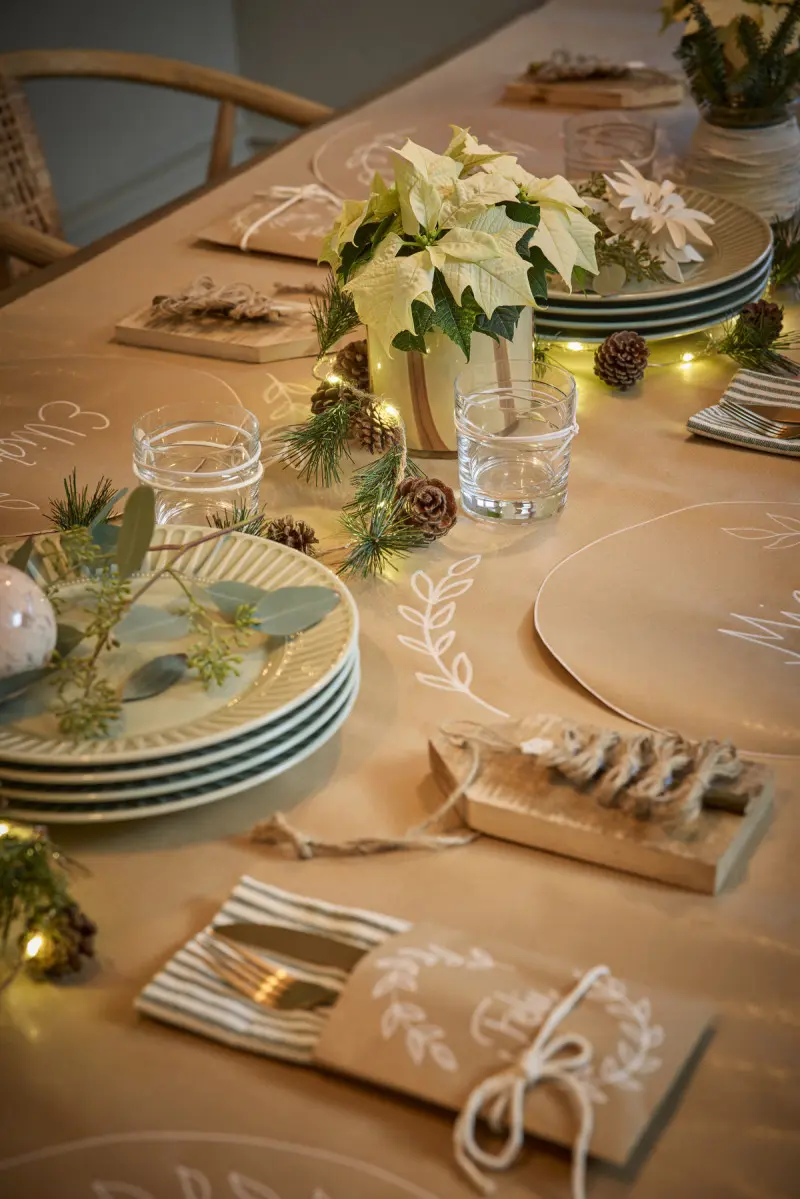 Świąteczny stół udekorowany w stylu skandynawskim z kremową poinsecją, gałązkami igliwia, szyszkami i ręcznie wykonanymi ozdobami z papieru.