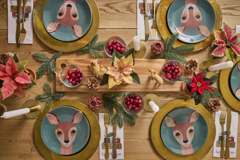Kolorowa, świąteczna dekoracja stołu z talerzami z motywem jelonka, czerwonymi i łososiowymi poinsecjami, żurawiną i gałązkami świerku.