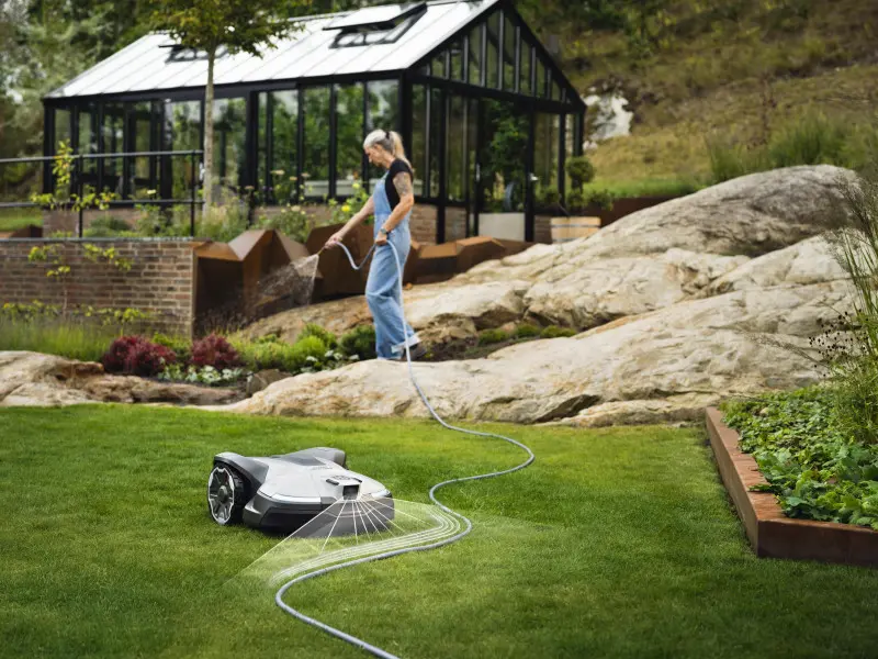 Robot koszący Husqvarna Automower na trawniku w ogrodzie, obok kobiety podlewającej rośliny przy nowoczesnej szklarni