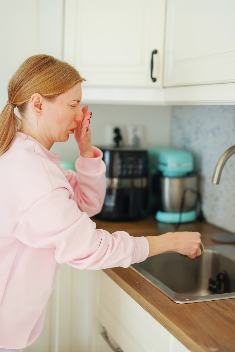 Kobieta zasłania nos nad zlewem, z którego wydobywa się brzydki zapach mimo czystości.