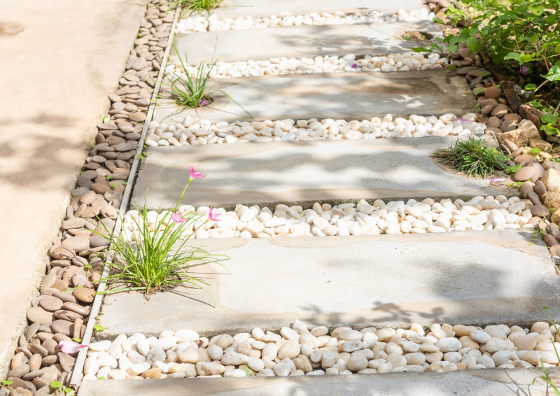 Prosta ścieżka z betonowych płyt wypełnionych białymi otoczakami i brązowymi kamieniami, z drobnymi roślinami między stopniami – elegancka nawierzchnia z kamieni w ogrodzie