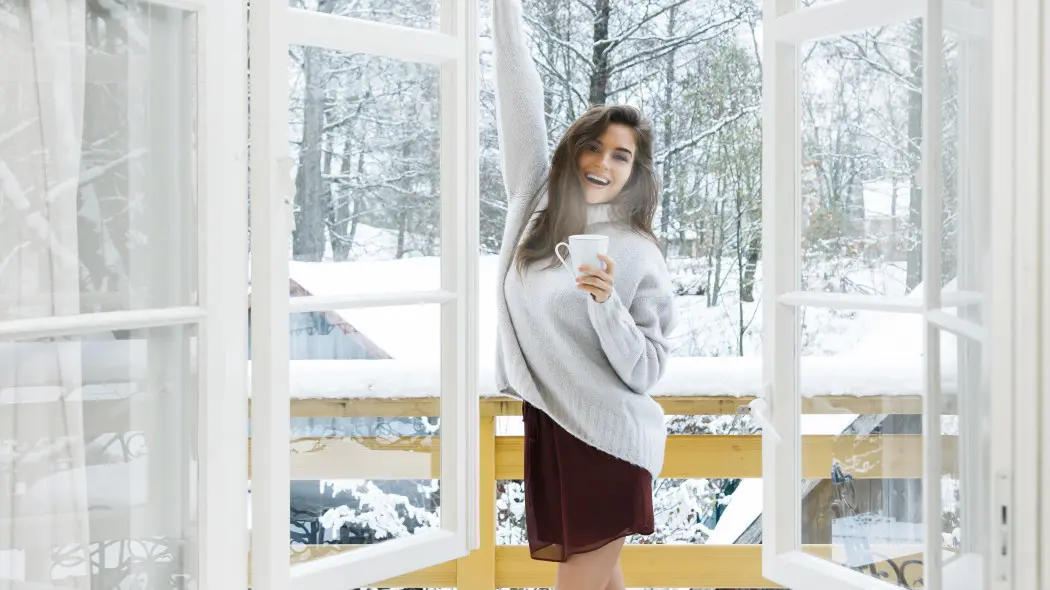 Kobieta z kubkiem przy otwartym oknie zimą – wietrzenie domu w sezonie grzewczym